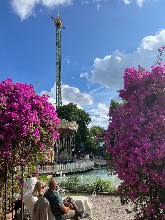People sitting under purple flowers with a tall amusement park ride in the background at Tivoli Gardens in Copenhagen 2025