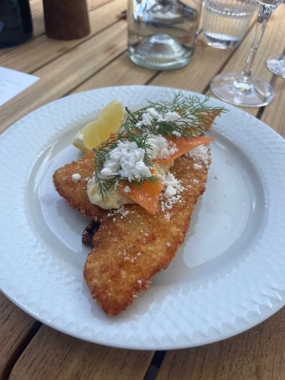 Dish of fried fish with lemon and herbs on a white plate, set on a wooden table. Lunch on Fritz Hansen during 3DD 2025 at Orangeriet