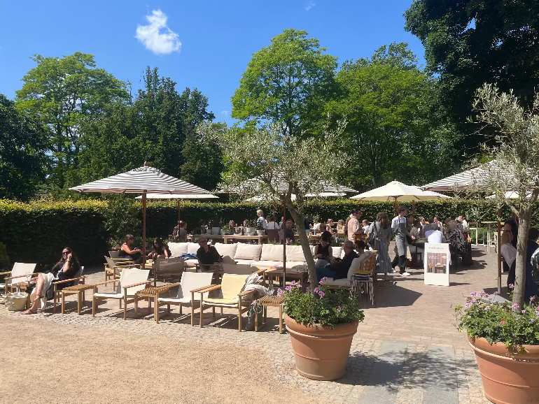 Orangiet restaurant in Copenhagen with Skagerak furniture. Outdoor patio area with people sitting at tables under umbrellas, surrounded by trees and potted plants.