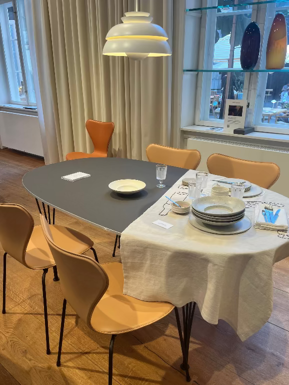 Dining table set with plates, glasses, and cutlery in a room with large windows and curtains. Fritz Hansen Tailored Series & chairs and super-elliptical dining table at their store in Copenhagen during 2025 3DD