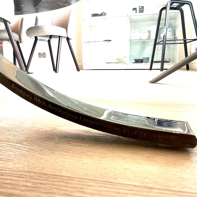 Curved metal object on a wooden surface with chairs and a table in the background. Knoll Barcelona Chair Bauhaus 100th Anniversary Limited Edition Black engraving on chrome leg