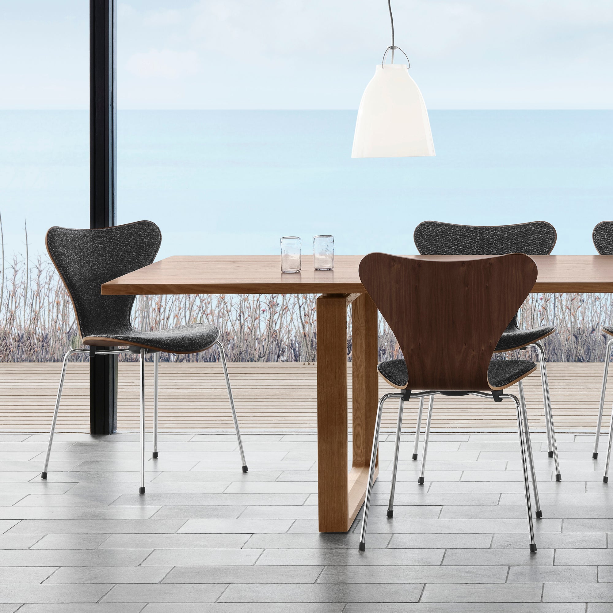 A modern dining table with a light wood top and thick central pedestal leg, surrounded by three chairs including one with a dark wood shell back and two gray upholstered chairs. The scene is set in a bright interior with a large window overlooking water and a wooden deck.