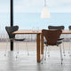 A modern dining table with a light wood top and thick central pedestal leg, surrounded by three chairs including one with a dark wood shell back and two gray upholstered chairs. The scene is set in a bright interior with a large window overlooking water and a wooden deck.