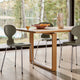 A modern dining table with a light oak wood top and sculptural metal legs, surrounded by three gray upholstered chairs with chrome legs, set in a bright room with large windows overlooking a lake.