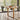 A modern dining table with a light oak wood top and sculptural metal legs, surrounded by three gray upholstered chairs with chrome legs, set in a bright room with large windows overlooking a lake.