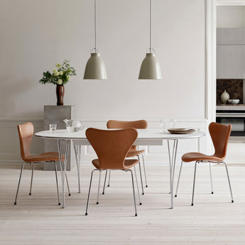 A dining set featuring four brown leather chairs with chrome legs around a white oval table, set in a bright, modern dining room with two pendant lights.