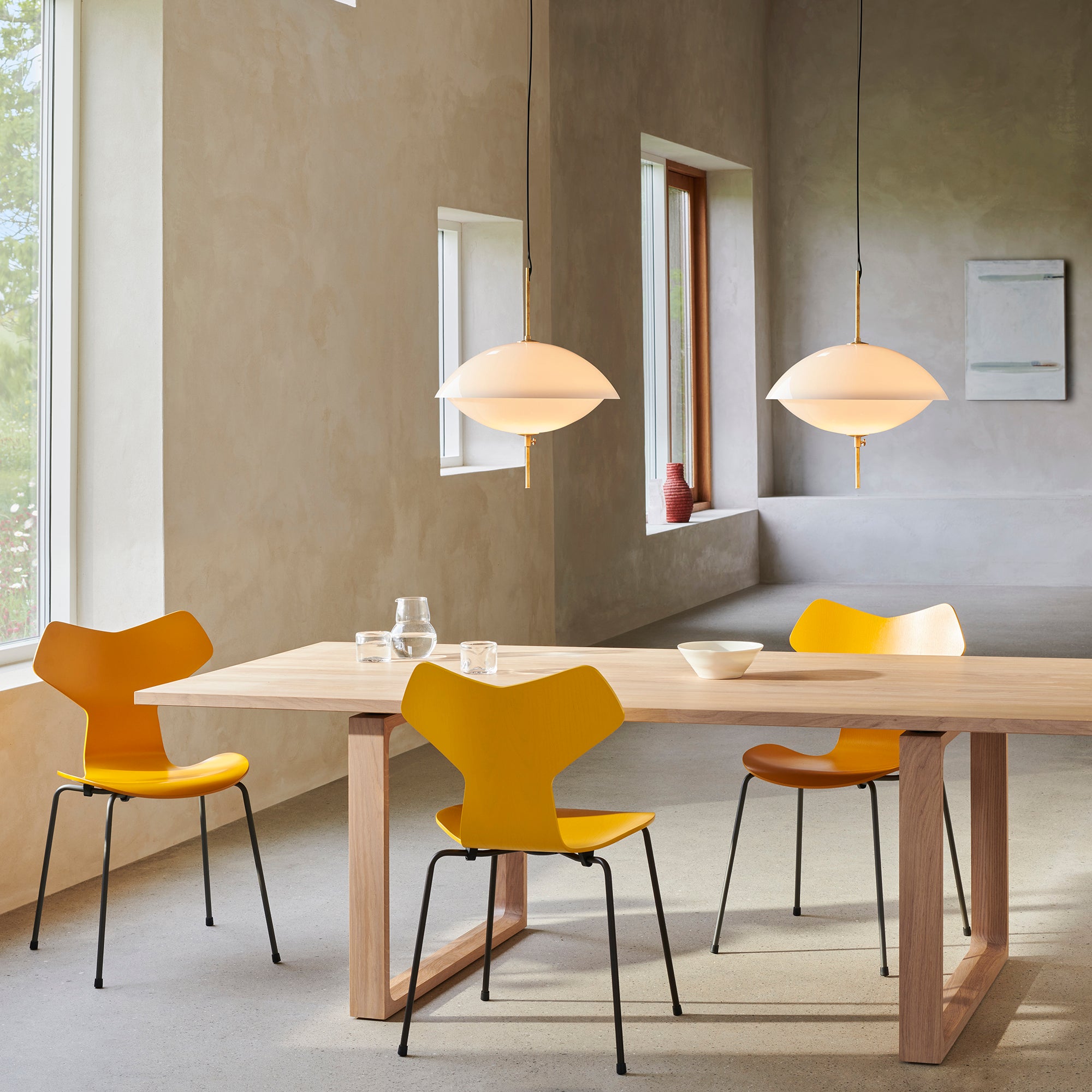A dining room scene with a light wood table and three modern chairs in bright yellow and orange, under two pendant lights in a minimalist interior with beige walls and windows.