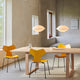 A dining room scene with a light wood table and three modern chairs in bright yellow and orange, under two pendant lights in a minimalist interior with beige walls and windows.