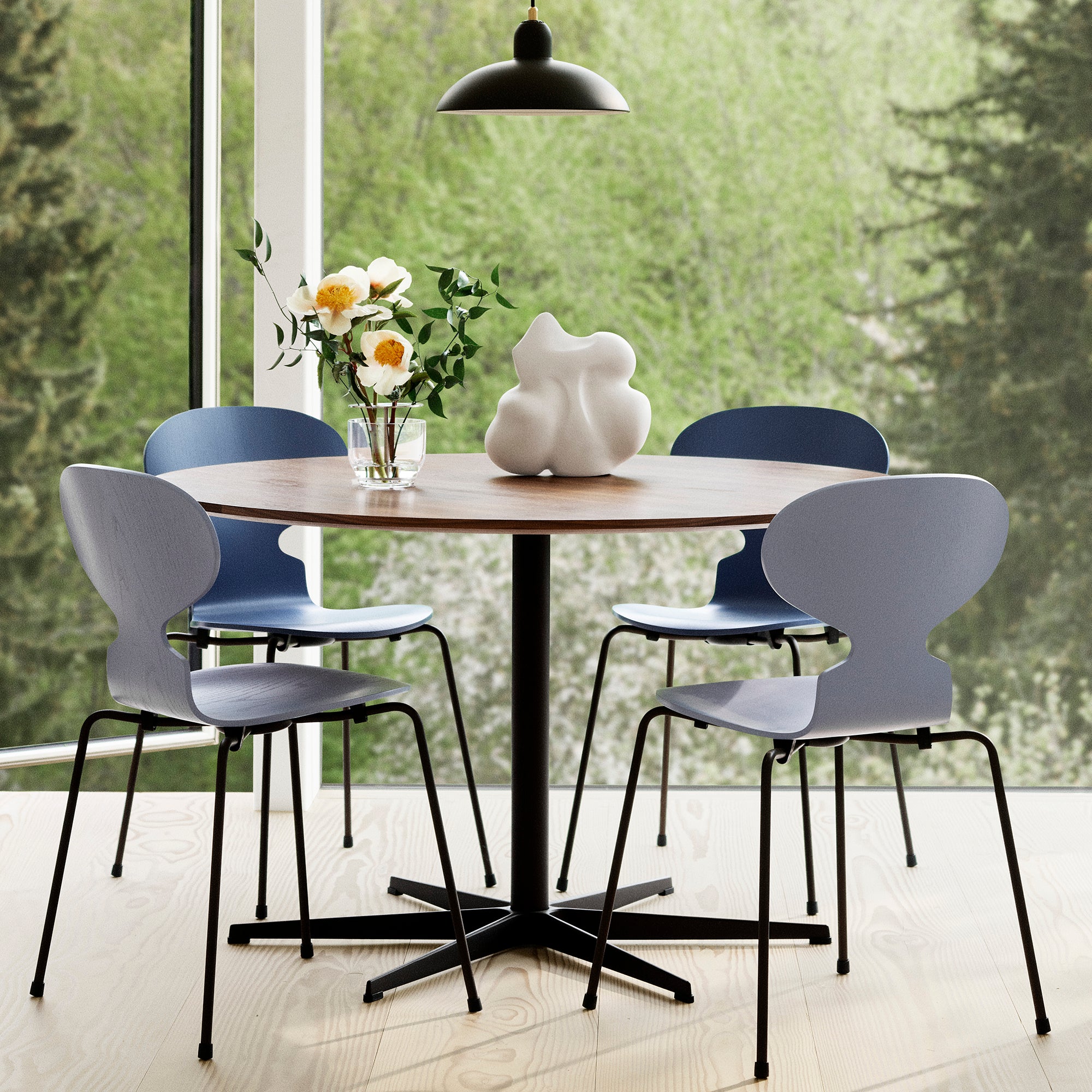 A round wooden dining table with a black metal pedestal base and four modern chairs with light gray molded seats and black legs, arranged in a bright room with large windows overlooking green trees. A vase of flowers and a white sculptural object sit on the table.