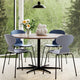 A round wooden dining table with a black metal pedestal base and four modern chairs with light gray molded seats and black legs, arranged in a bright room with large windows overlooking green trees. A vase of flowers and a white sculptural object sit on the table.