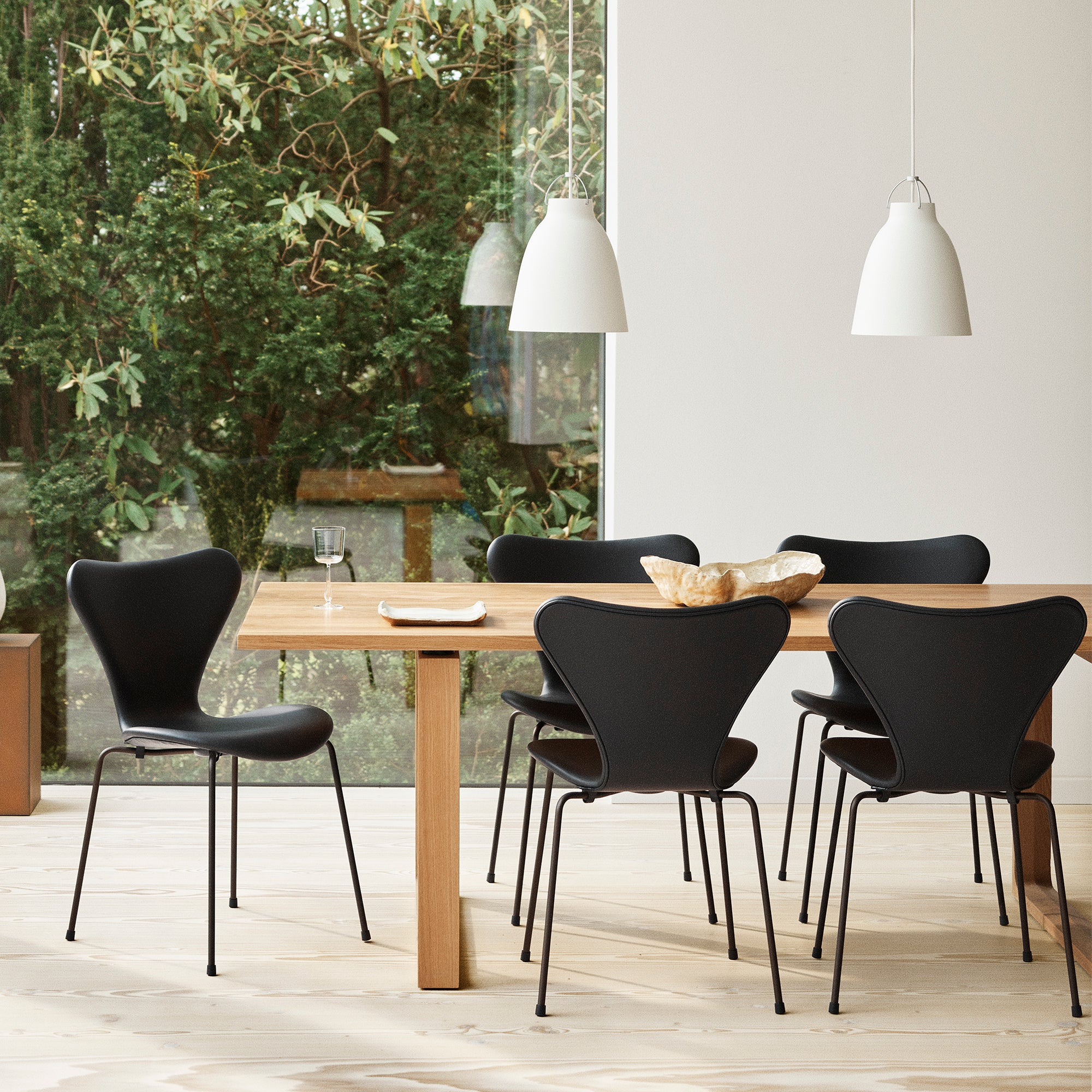 A dining room scene with a light wood table and four black chairs, including a prominent sculptural black chair in the foreground. White pendant lights hang above, and a large window behind shows green foliage outside.