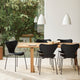 A dining room scene with a light wood table and four black chairs, including a prominent sculptural black chair in the foreground. White pendant lights hang above, and a large window behind shows green foliage outside.