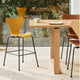 A modern kitchen or dining area scene with a light wood table and multiple bar stools. The most prominent product is a tall stool with a bright yellow molded seat and back, black metal legs, and a footrest.