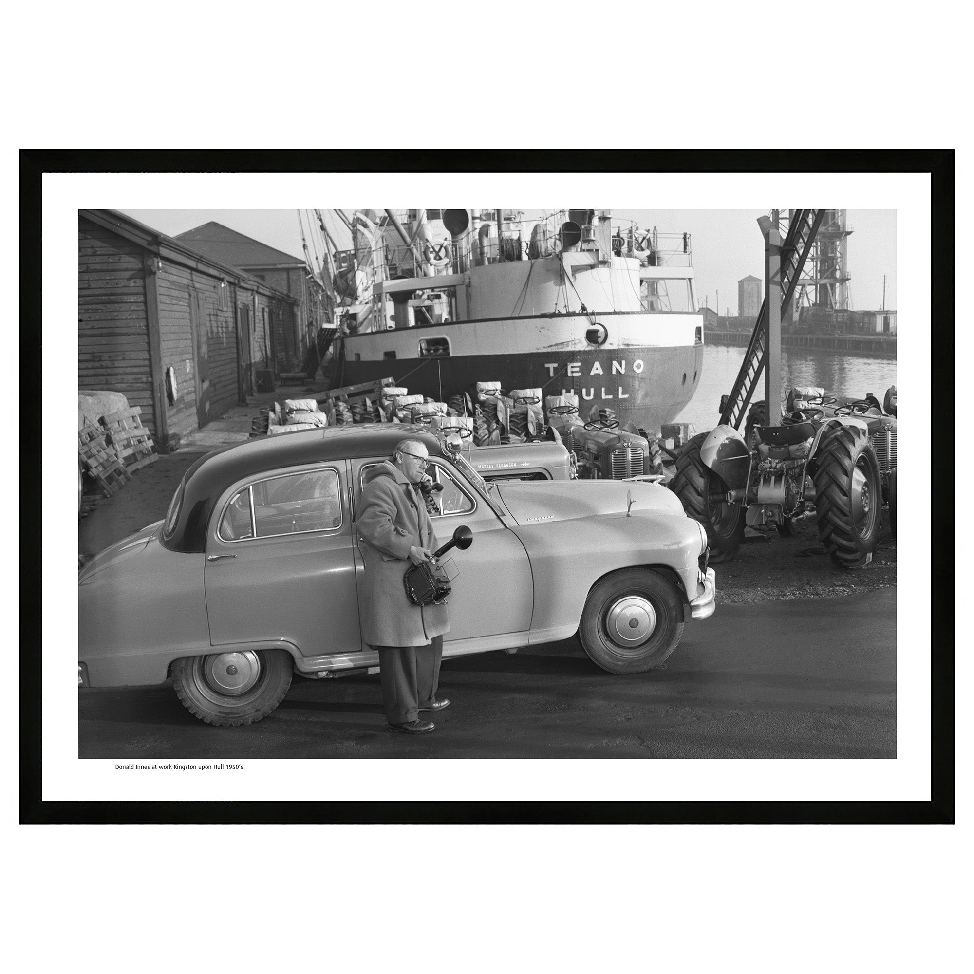 Innes Heritage Donald Innes At Work 1950 A2 Framed Art Print in Black frame Standard Vanguard car with man on car phone and holding a vintage camera on a dockside in hull with Teano Hull trawler and massey ferguson tractors on quayside waiting to be loaded up for export. historical image back from 1950