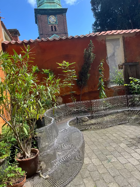 Decorative Pantonova wire bench in a garden setting with a building and clock tower in the background. Montana store at 3 days of design 2025 Copenhagen