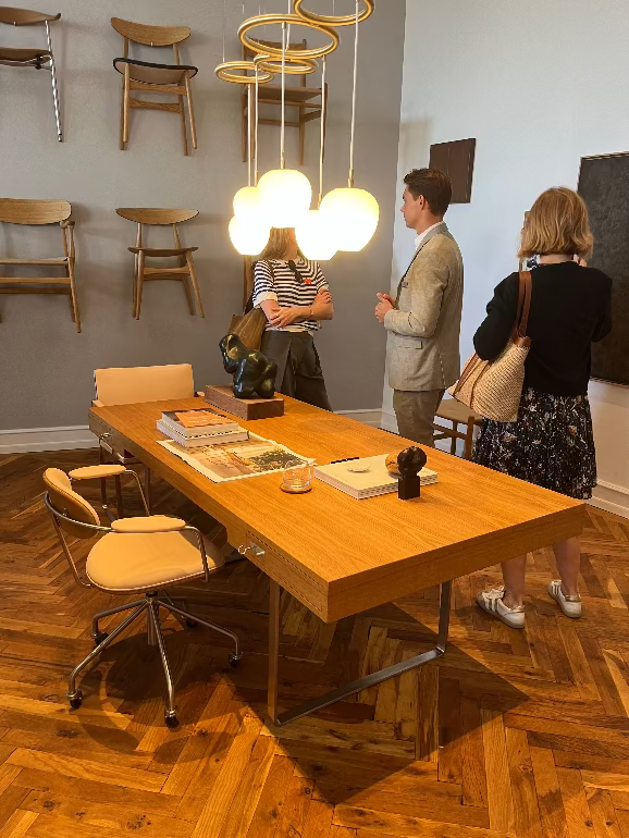 People interacting around a wooden table in a modern interior setting with chairs and hanging lights. Carl Hansen Copenhagen showroom 3DD 2025. Hans Wegner CH110 desk and chair.