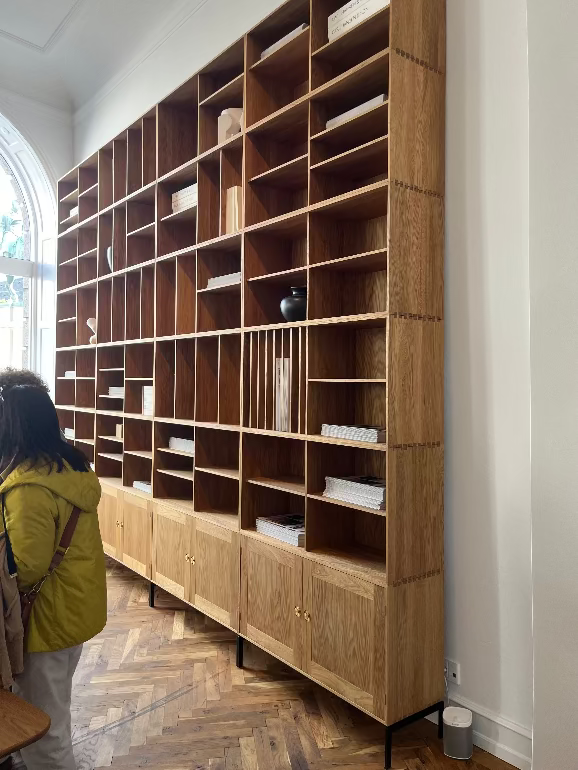 Large wooden bookshelf in a room with a person looking at it.Carl Hansen Copenhagen showroom 3DD 2025. Ole Wanscher shelving