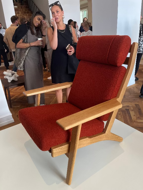 Red armchair with wooden frame on a white platform, people in the background. Carl Hansen Copenhagen showroom 3DD 2025
