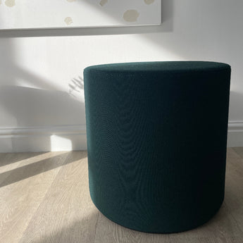 Dark green cylindrical stool on a light wooden floor with a white wall in the background. Vitra's visiona upholstered stool by verner panton in volo green fabric, on display at innes gallery in hessle square, uk