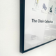 A close-up of a framed wall art print titled 'The Chair Collection' hanging on a white textured wall. The print features minimalist chair illustrations and text, enclosed in a thin dark frame.
