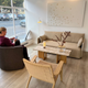 A modern living room setup featuring a beige fabric sofa, a marble-top coffee table, a woven wood armchair, and a dark brown armchair where a woman is seated by a large window. Audo Offset 2 seater sofa in Boucle 02. CH25 oak lounge & brown fabric Audo Eave Seamline Lounge Chair.