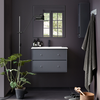 A modern bathroom interior with a floating gray vanity, white sink top, dark charcoal walls and floor, a mirror, floor lamp, hanging towel, and a potted plant.
