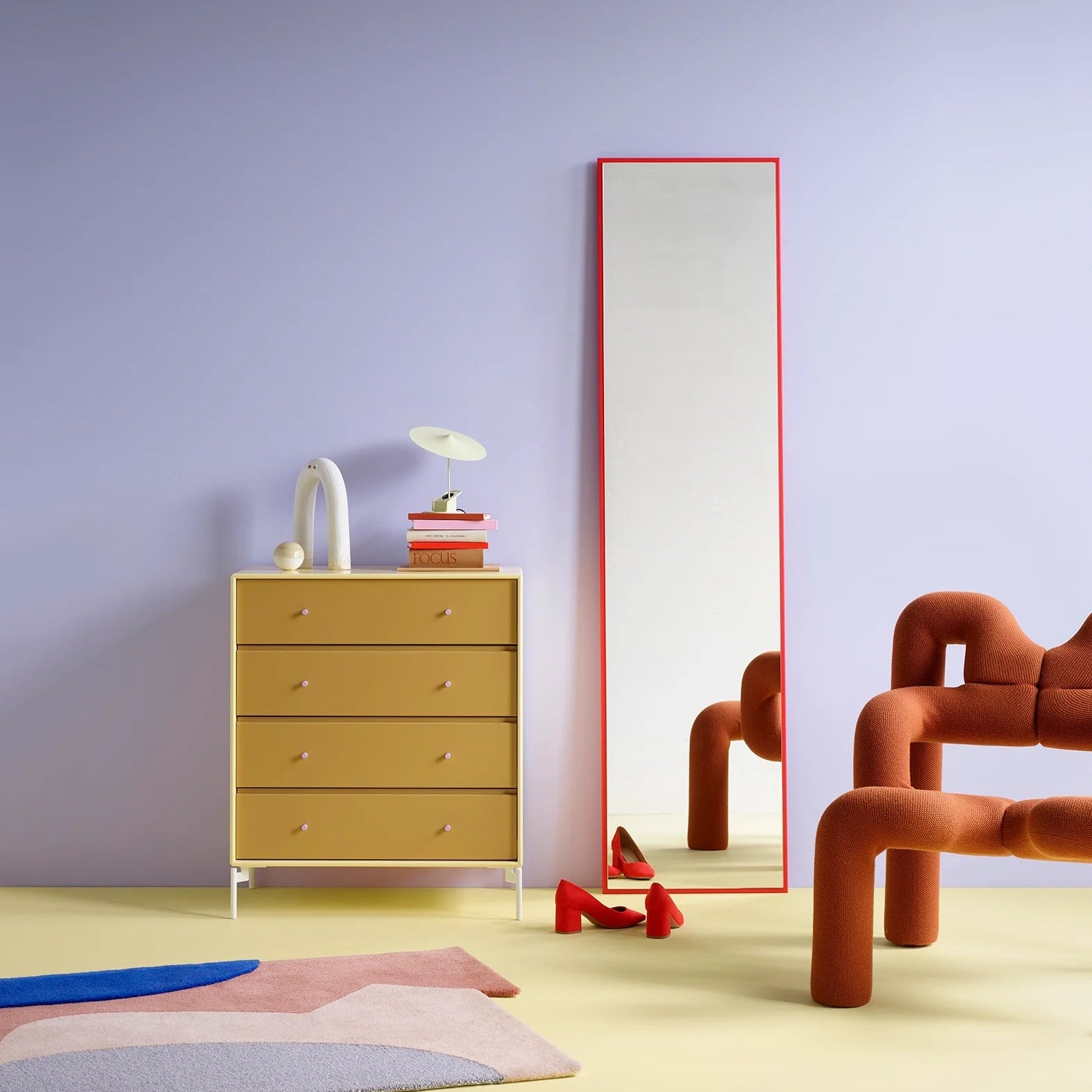 A tall, narrow floor mirror with a thin red frame stands against a lavender wall in a stylized room. To the left is a mustard yellow four-drawer dresser with small knobs, topped with a white vase, a small lamp, and stacked books.
