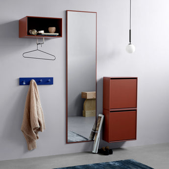 A modern interior scene showing a tall floor mirror with a reddish-brown frame, a matching wall-mounted open shelf, a blue coat rack with a beige sweater hanging from it, and a two-door shoe cabinet in the same reddish-brown finish. A dark blue rug is on the floor, and a pendant light hangs above.