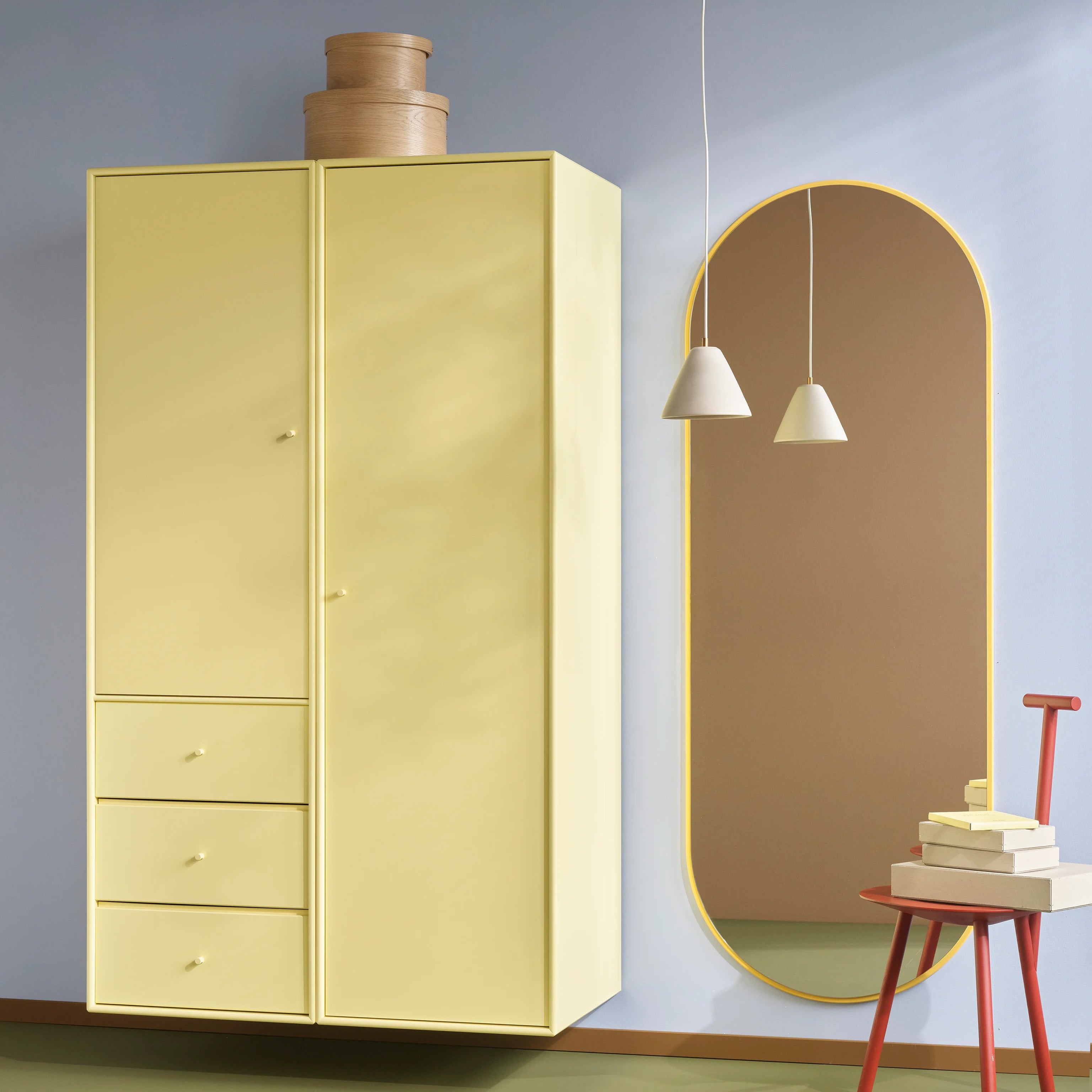 A pale yellow wooden wardrobe with five drawers on the left, shown in a styled room next to an arched mirror, pendant lights, a small red stool, and a rug.
